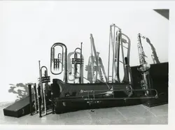 Uma fotografia em preto e branco exibindo uma coleção de instrumentos musicais de sopro, incluindo trompetes, trombones e um saxofone, dispostos sobre ou próximos a seus estojos. A imagem sugere um contexto de banda musical ou fanfarra, possivelmente em uma cidade pequena como Videira, com uma estética que remete à metade do século XX.