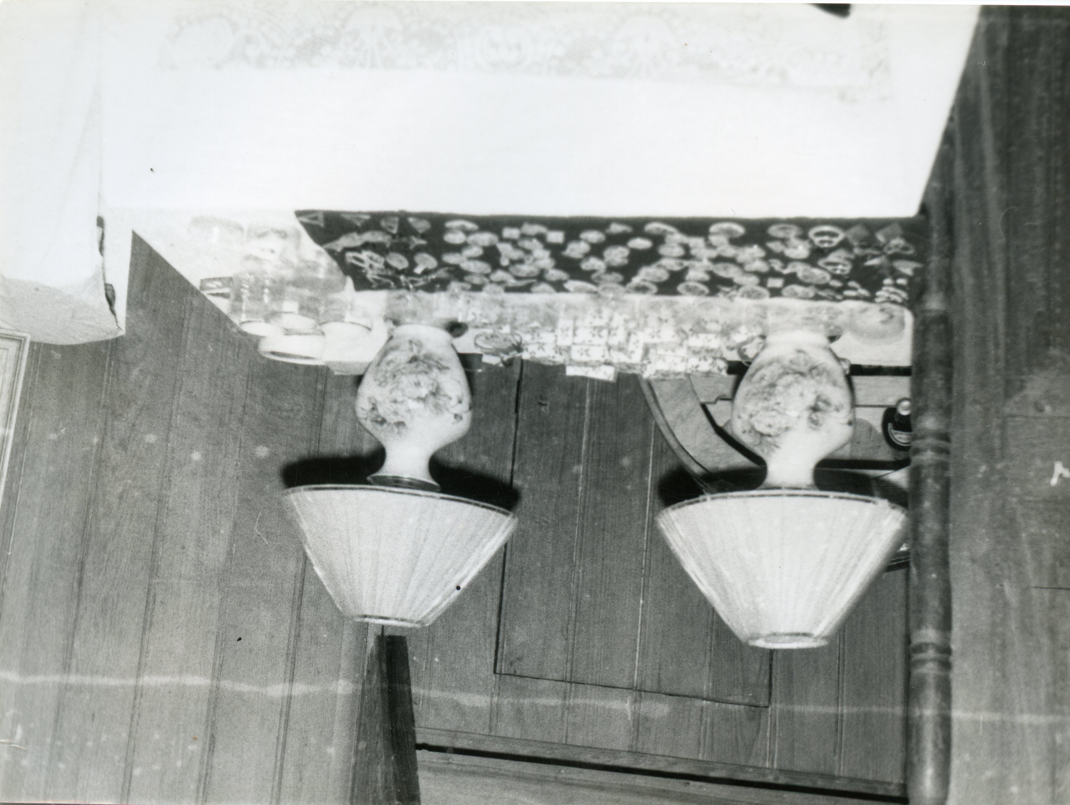Fotografia em preto e branco capturando um detalhe de um ambiente doméstico, possivelmente uma mesa ou aparador com dois abajures de estilo clássico. A imagem apresenta uma perspectiva inusitada, com os objetos dispostos sobre uma superfície de madeira, sugerindo um cenário residencial típico de meados do século XX.