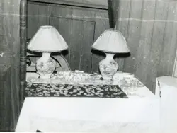 Fotografia em preto e branco capturando um detalhe de um ambiente doméstico, possivelmente uma mesa ou aparador com dois abajures de estilo clássico. A imagem apresenta uma perspectiva inusitada, com os objetos dispostos sobre uma superfície de madeira, sugerindo um cenário residencial típico de meados do século XX.