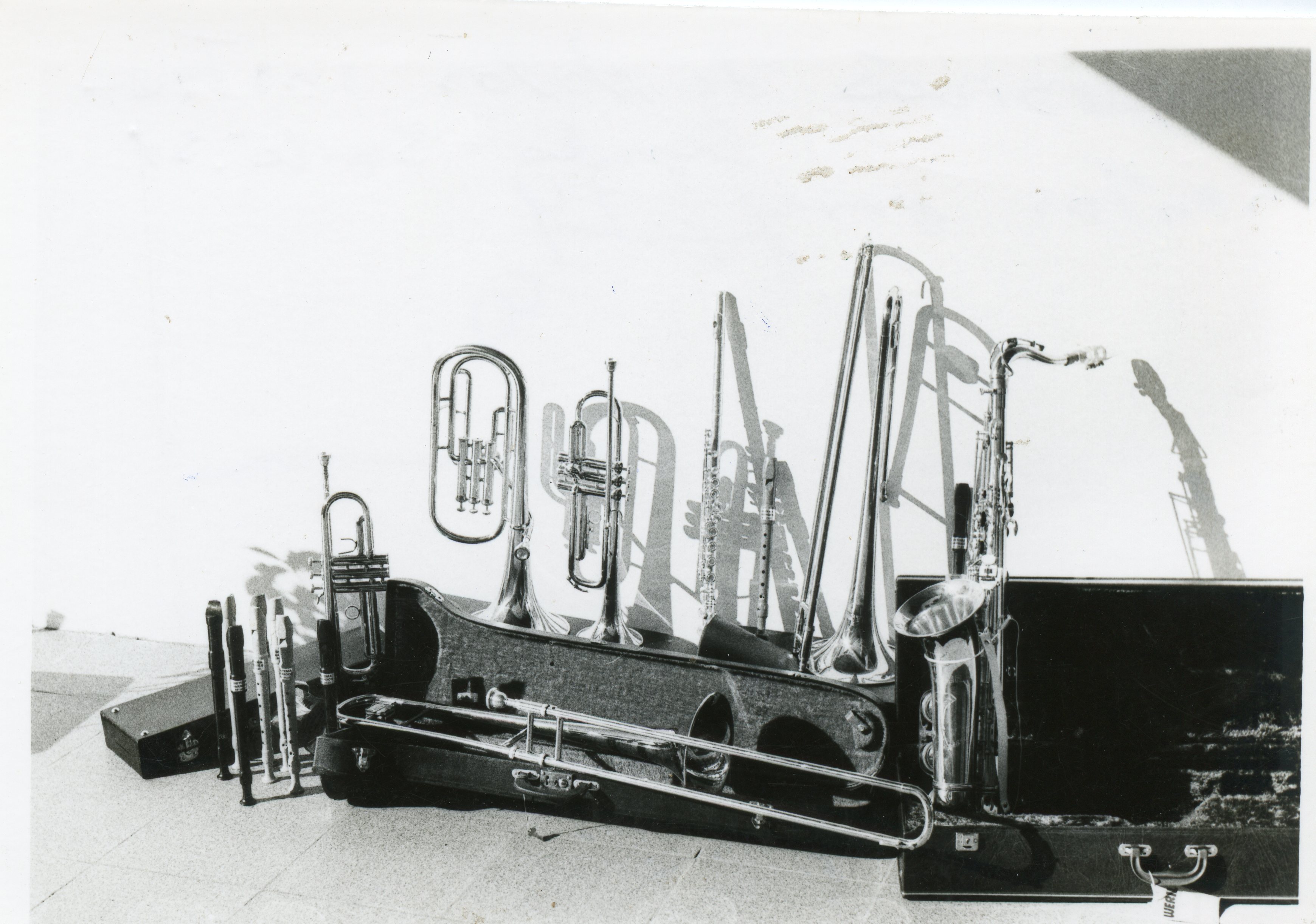 Uma fotografia em preto e branco exibindo uma coleção de instrumentos musicais de sopro, incluindo trompetes, trombones, flautas e um saxofone, dispostos sobre o chão. A iluminação lateral cria sombras marcantes na parede branca ao fundo, sugerindo um ambiente de ensaio ou preparação para uma apresentação musical.
