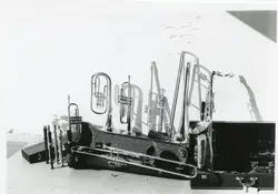 Uma fotografia em preto e branco exibindo uma coleção de instrumentos musicais de sopro, incluindo trompetes, trombones, flautas e um saxofone, dispostos sobre o chão. A iluminação lateral cria sombras marcantes na parede branca ao fundo, sugerindo um ambiente de ensaio ou preparação para uma apresentação musical.