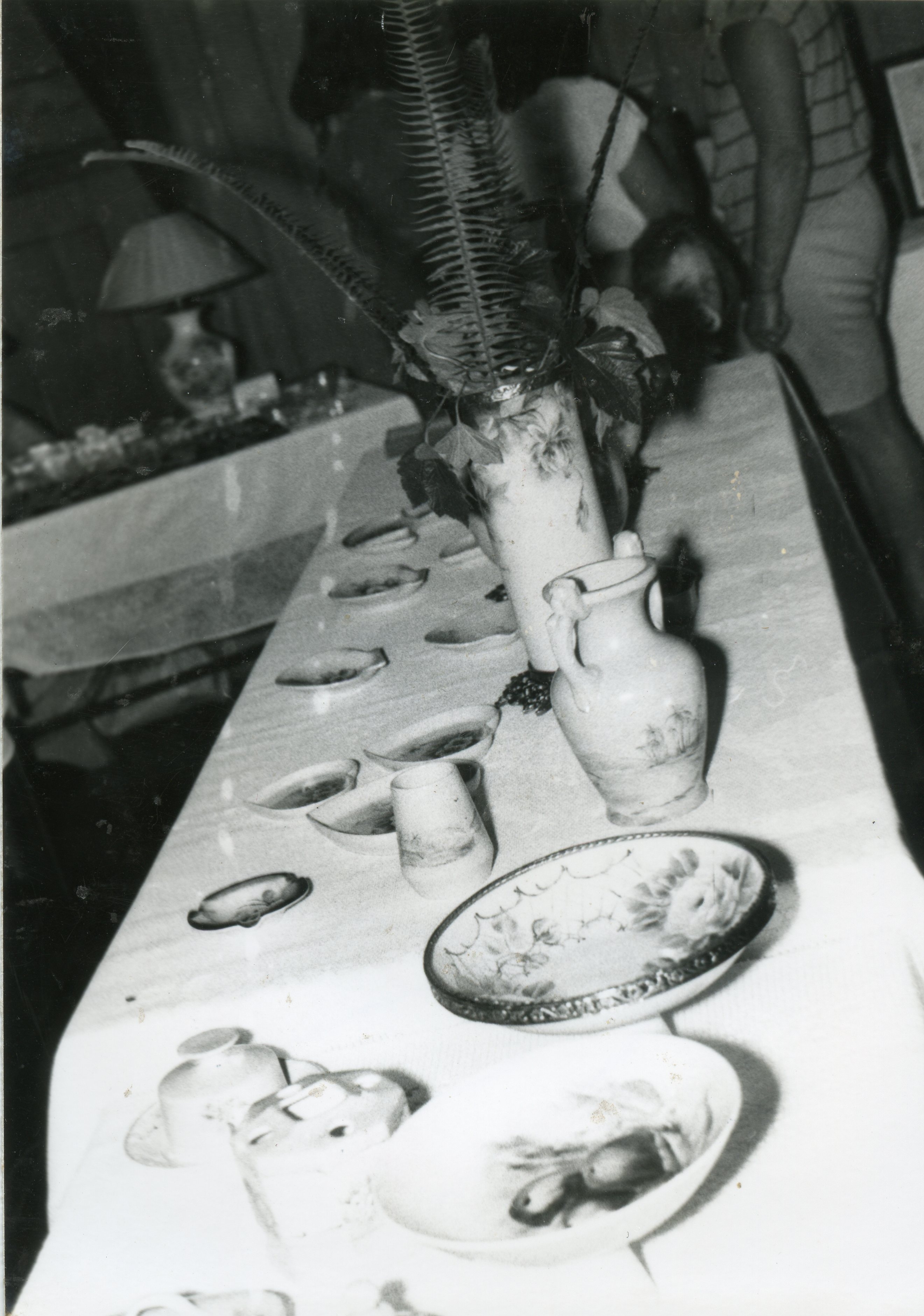 Fotografia em preto e branco exibindo uma mesa posta com diversos itens de louça decorada, incluindo vasos, pratos e tigelas. O cenário sugere um evento social ou uma exposição doméstica, possivelmente ocorrida em meados do século XX, com pessoas parcialmente visíveis ao fundo.