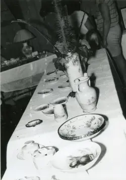 Fotografia em preto e branco exibindo uma mesa posta com diversos itens de louça decorada, incluindo vasos, pratos e tigelas. O cenário sugere um evento social ou uma exposição doméstica, possivelmente ocorrida em meados do século XX, com pessoas parcialmente visíveis ao fundo.