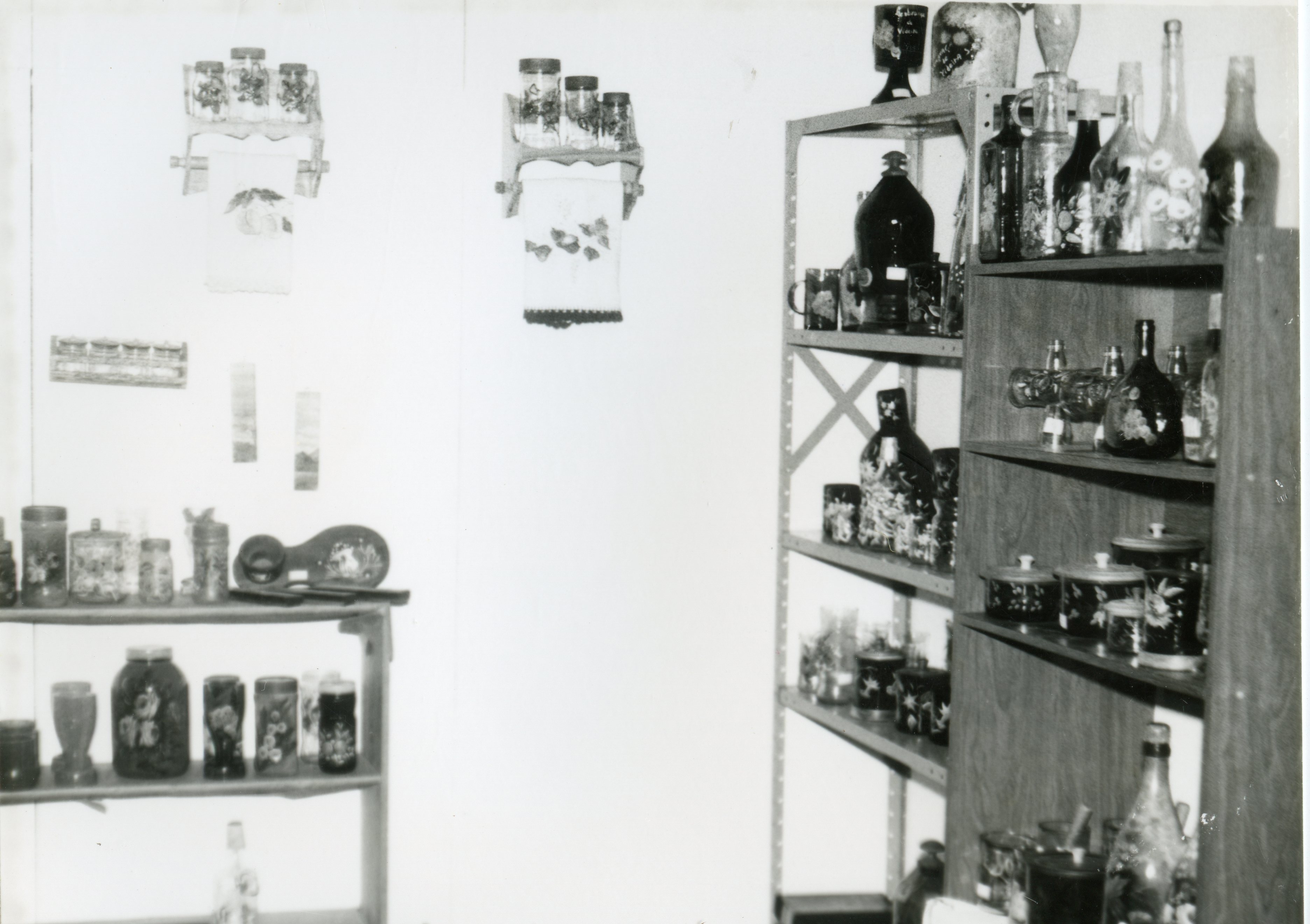 Fotografia em preto e branco de um interior doméstico, possivelmente uma despensa ou cozinha, exibindo prateleiras repletas de potes de vidro decorados e garrafas. O ambiente apresenta elementos de artesanato típico de meados do século XX, sugerindo um contexto de organização doméstica em uma residência brasileira.
