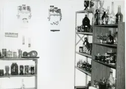 Fotografia em preto e branco de um interior doméstico, possivelmente uma despensa ou cozinha, exibindo prateleiras repletas de potes de vidro decorados e garrafas. O ambiente apresenta elementos de artesanato típico de meados do século XX, sugerindo um contexto de organização doméstica em uma residência brasileira.