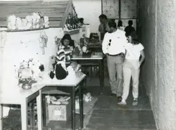 Fotografia em preto e branco registrando um ambiente interno, possivelmente uma feira de artesanato ou exposição escolar, com pessoas circulando entre mesas com objetos decorativos. O estilo das vestimentas, como as calças de cintura alta e camisetas, sugere um contexto da década de 1980 ou início de 1990.