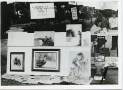 Uma fotografia em preto e branco que mostra uma exposição de arte ao ar livre, possivelmente em uma praça pública. Várias pinturas estão dispostas sobre um suporte, acompanhadas por cartazes com frases reflexivas e uma pessoa sentada à mesa, sugerindo um evento cultural ou feira de artesanato de meados do século XX.