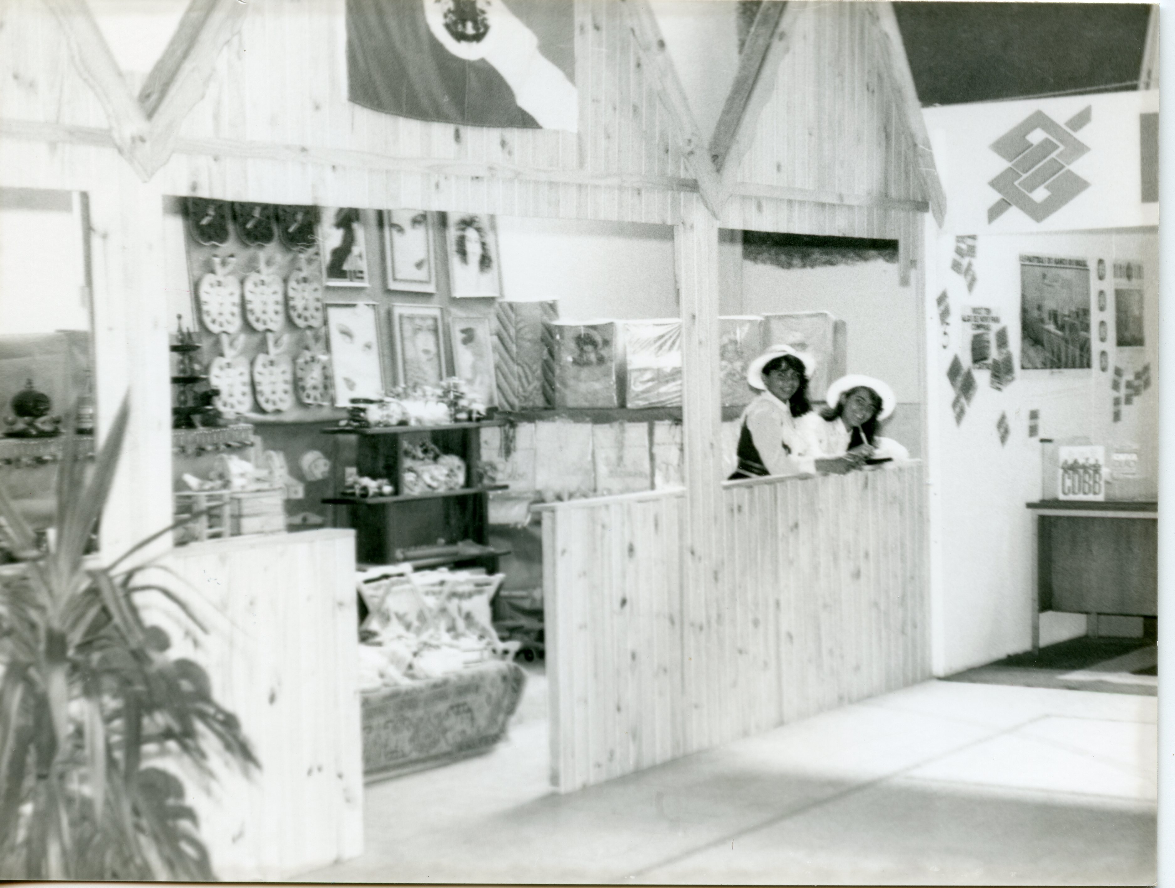 Fotografia em preto e branco mostrando um estande de exposição, possivelmente em uma feira comercial ou evento público. Duas mulheres com chapéus estão atrás de um balcão de madeira, com diversos produtos decorativos e o logotipo do Banco do Brasil visível ao fundo, sugerindo um contexto institucional ou promocional das décadas de 1970 ou 1980.