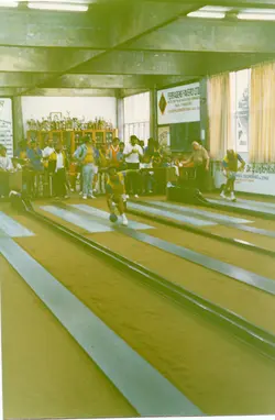 Uma fotografia colorida que retrata uma partida de bolão em um clube social, provavelmente em uma cidade do interior do Brasil como Videira. O ambiente apresenta pistas de madeira, jogadores uniformizados e uma plateia ao fundo, com elementos decorativos como troféus e anúncios comerciais nas paredes, sugerindo um evento esportivo comunitário da década de 1980.