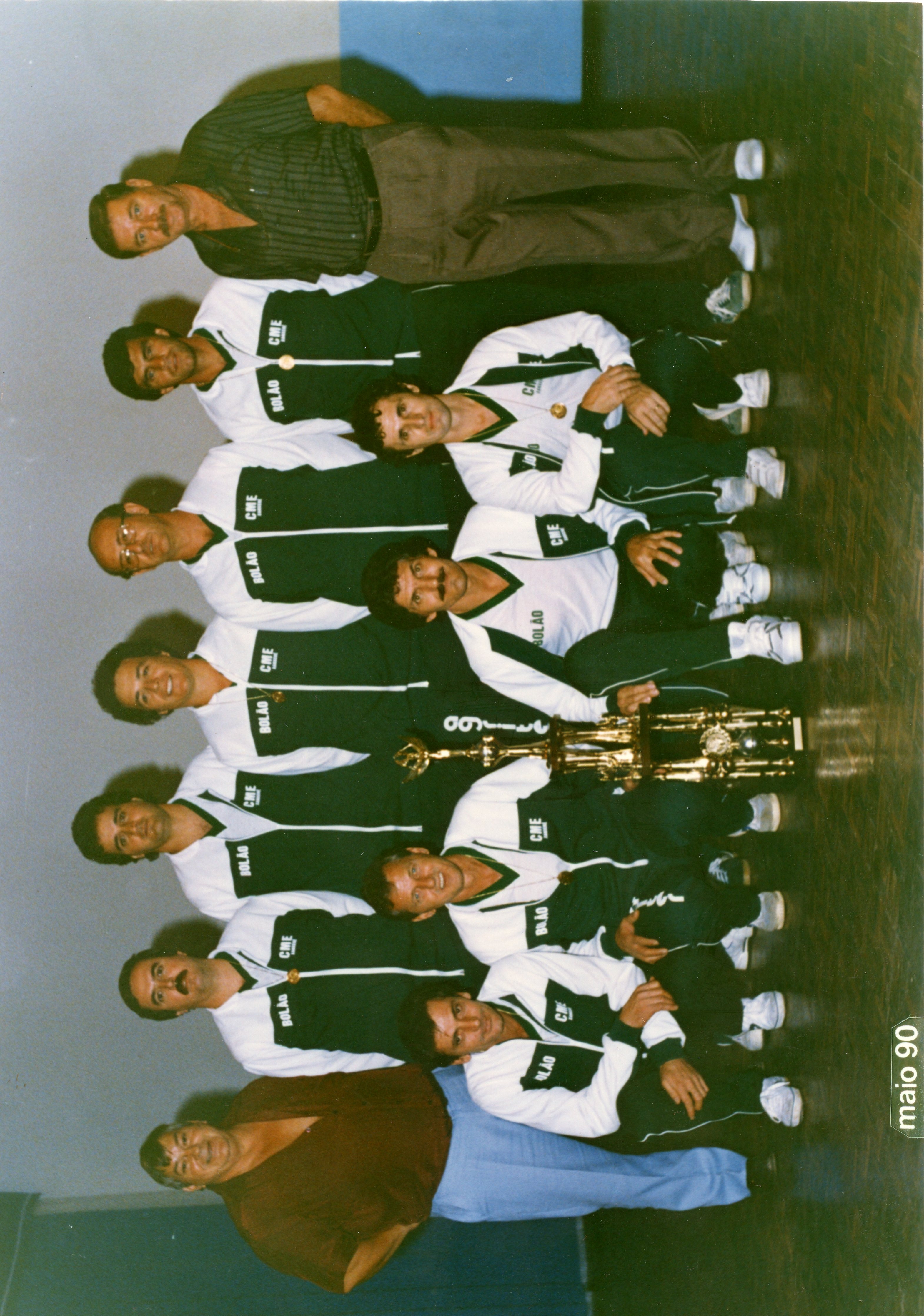 Fotografia de grupo de uma equipe de bolão, composta por homens vestindo agasalhos esportivos padronizados com a inscrição 'CME' e 'BOLÃO'. O grupo posa com um troféu, sugerindo uma celebração de vitória esportiva em um ambiente interno, datada de maio de 1990.