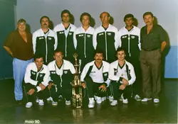 Fotografia de grupo de uma equipe de bolão, composta por homens vestindo agasalhos esportivos padronizados com a inscrição 'CME' e 'BOLÃO'. O grupo posa com um troféu, sugerindo uma celebração de vitória esportiva em um ambiente interno, datada de maio de 1990.