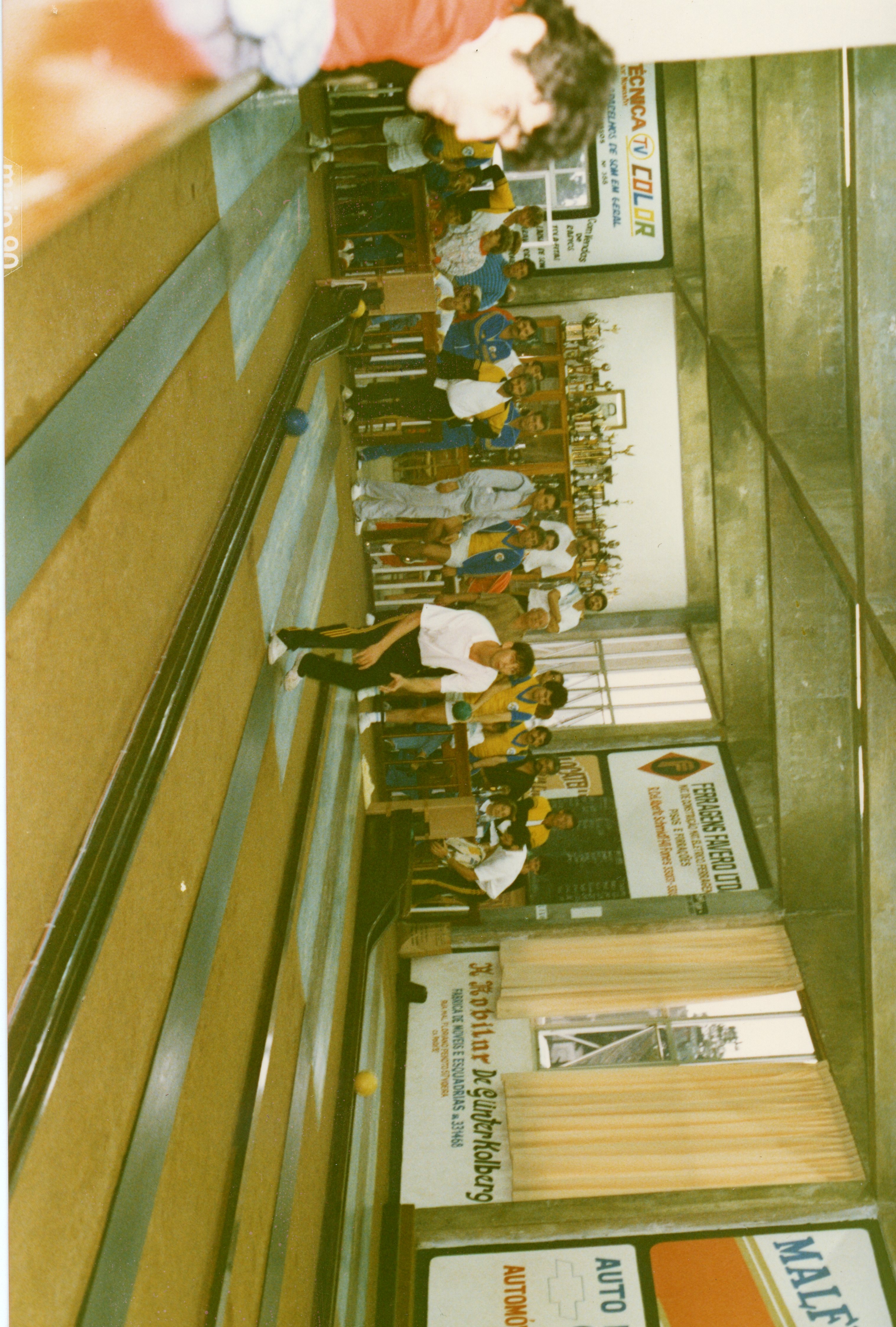 Uma fotografia de um salão de boliche com várias pessoas praticando o esporte, possivelmente em um clube social ou recreativo. O ambiente apresenta diversos anúncios comerciais nas paredes, típicos de estabelecimentos locais brasileiros, sugerindo uma cena da década de 1980 ou 1990.