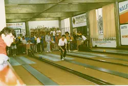 Uma fotografia de um salão de boliche com várias pessoas praticando o esporte, possivelmente em um clube social ou recreativo. O ambiente apresenta diversos anúncios comerciais nas paredes, típicos de estabelecimentos locais brasileiros, sugerindo uma cena da década de 1980 ou 1990.