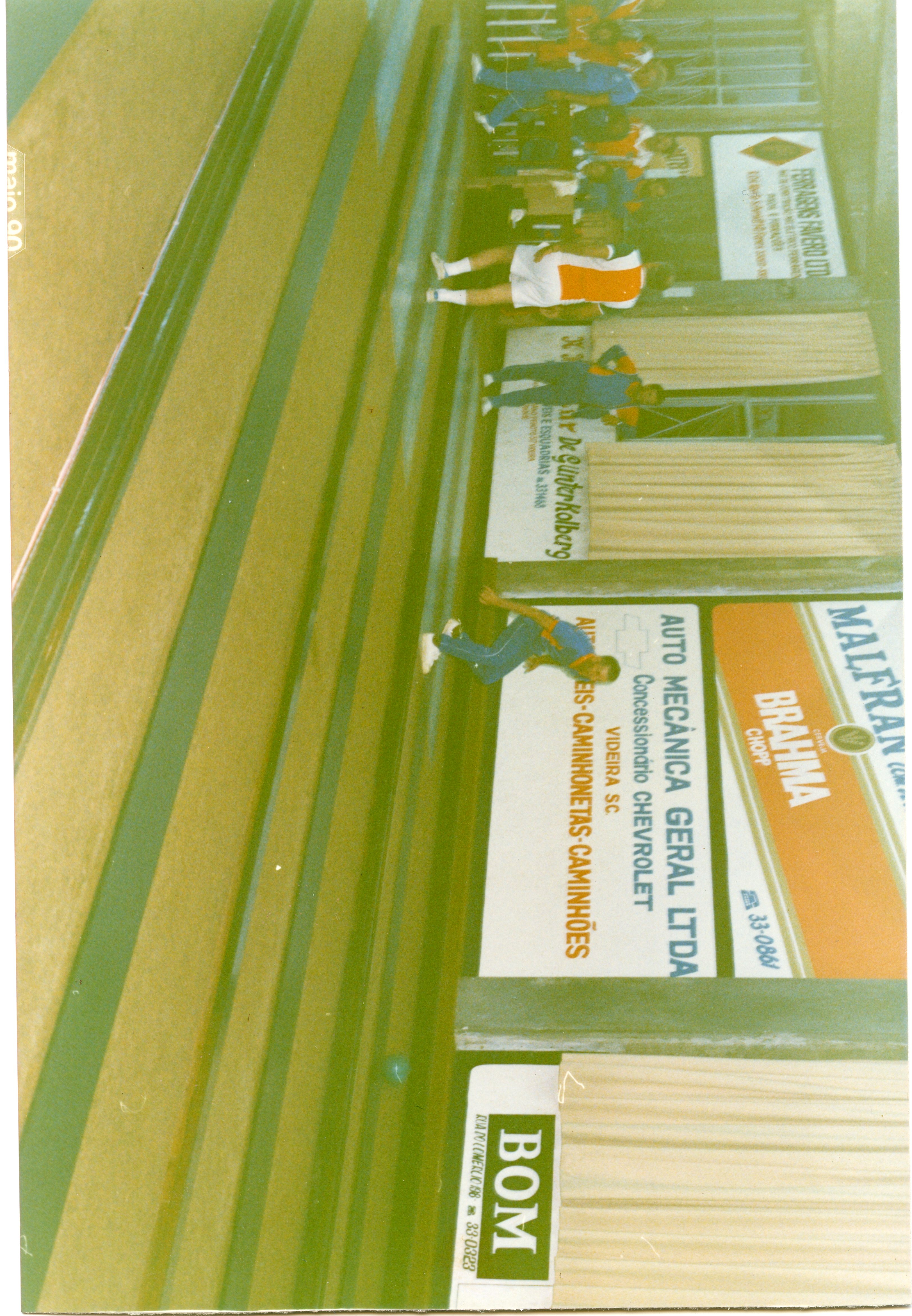 Fotografia de um ambiente esportivo interno, possivelmente uma cancha de bocha ou boliche, com pessoas vestindo roupas esportivas da década de 1990. O local apresenta diversos anúncios publicitários de empresas locais de Videira, SC, como concessionárias e comércios, indicando um evento ou prática recreativa comunitária.