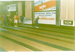 Fotografia de um ambiente esportivo interno, possivelmente uma cancha de bocha ou boliche, com pessoas vestindo roupas esportivas da década de 1990. O local apresenta diversos anúncios publicitários de empresas locais de Videira, SC, como concessionárias e comércios, indicando um evento ou prática recreativa comunitária.
