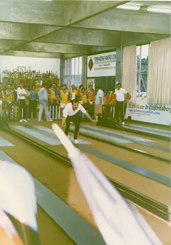 Uma fotografia colorida registrando uma partida de bolão em um ambiente fechado, provavelmente um clube social em Videira, Santa Catarina. O registro mostra um jogador em movimento na pista, cercado por espectadores e troféus ao fundo, com banners publicitários locais visíveis, datado de maio de 1990.