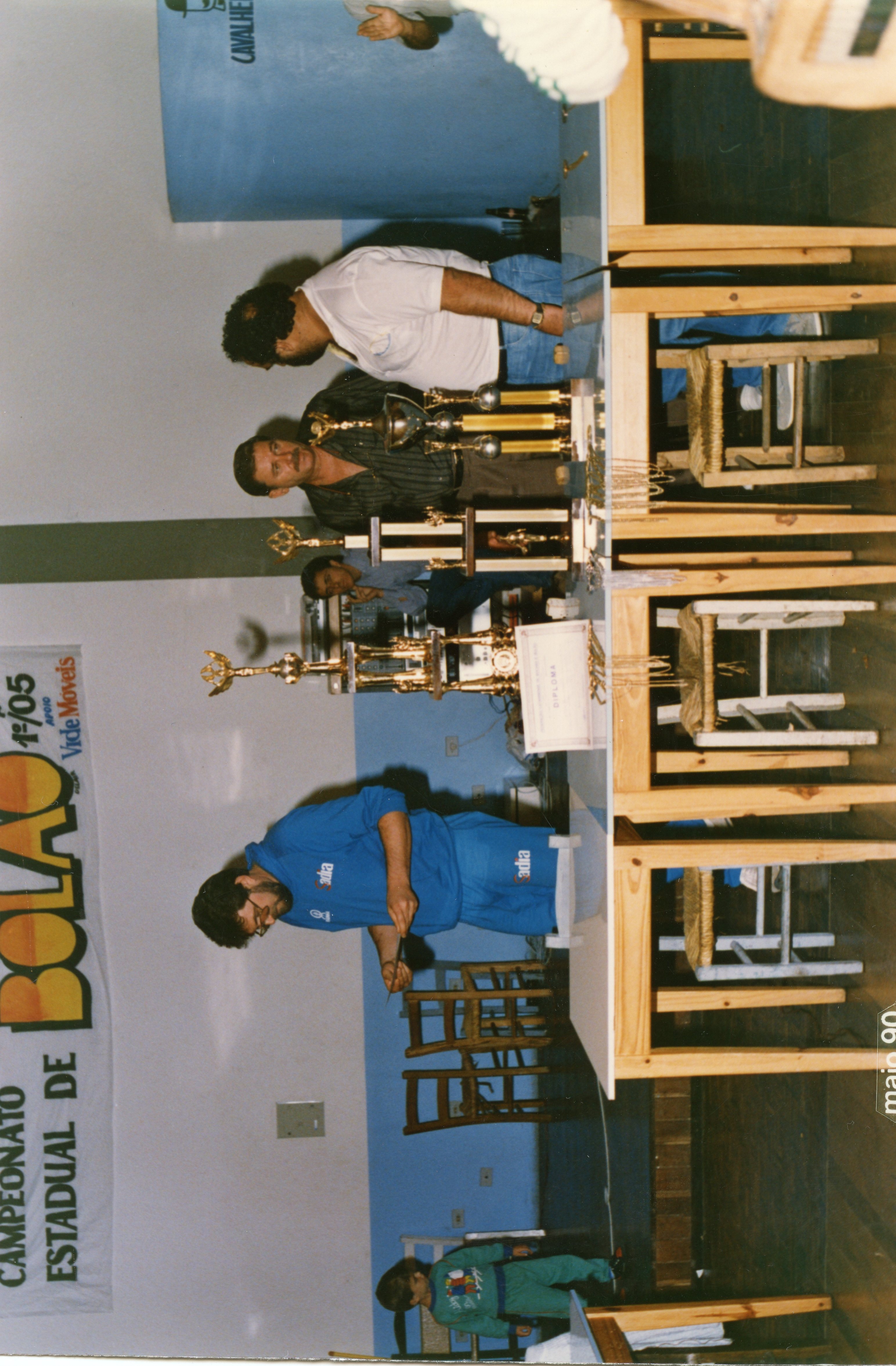 Fotografia colorida de uma cerimônia de premiação de um campeonato de bolão, datada de maio de 1990. Homens estão organizando troféus sobre uma mesa em um ambiente interno, com um banner promocional visível ao lado, sugerindo um evento esportivo regional em Videira.