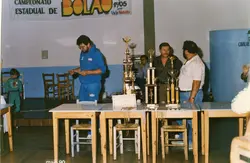 Fotografia colorida de uma cerimônia de premiação de um campeonato de bolão, datada de maio de 1990. Homens estão organizando troféus sobre uma mesa em um ambiente interno, com um banner promocional visível ao lado, sugerindo um evento esportivo regional em Videira.