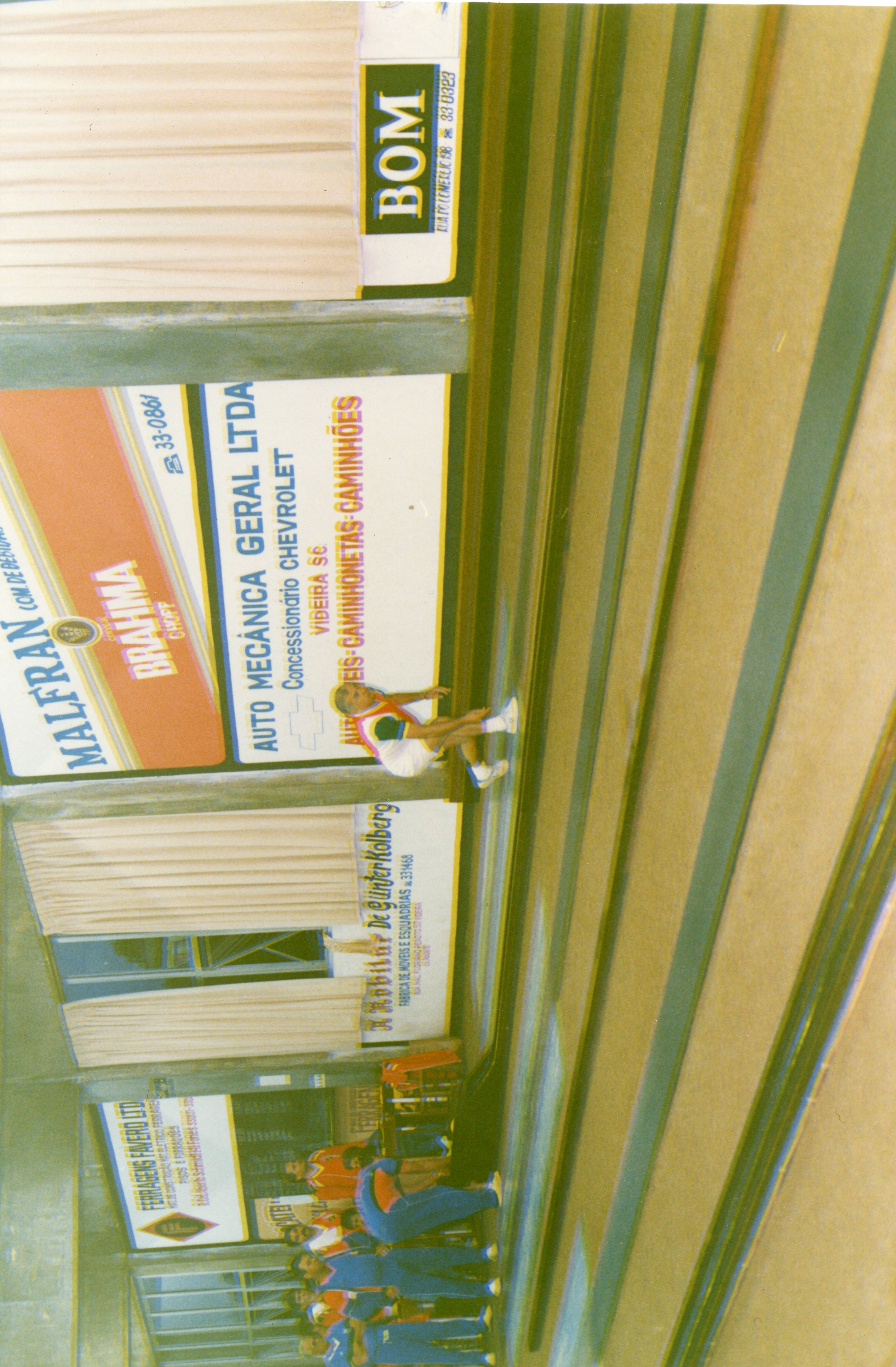 Uma fotografia colorida, orientada verticalmente, que mostra uma arquibancada ou área de eventos em Videira, Santa Catarina, com diversas placas publicitárias de empresas locais da época. A imagem exibe pessoas sentadas ou em pé na estrutura, sugerindo um evento esportivo ou comunitário, possivelmente nas décadas de 1980 ou 1990.