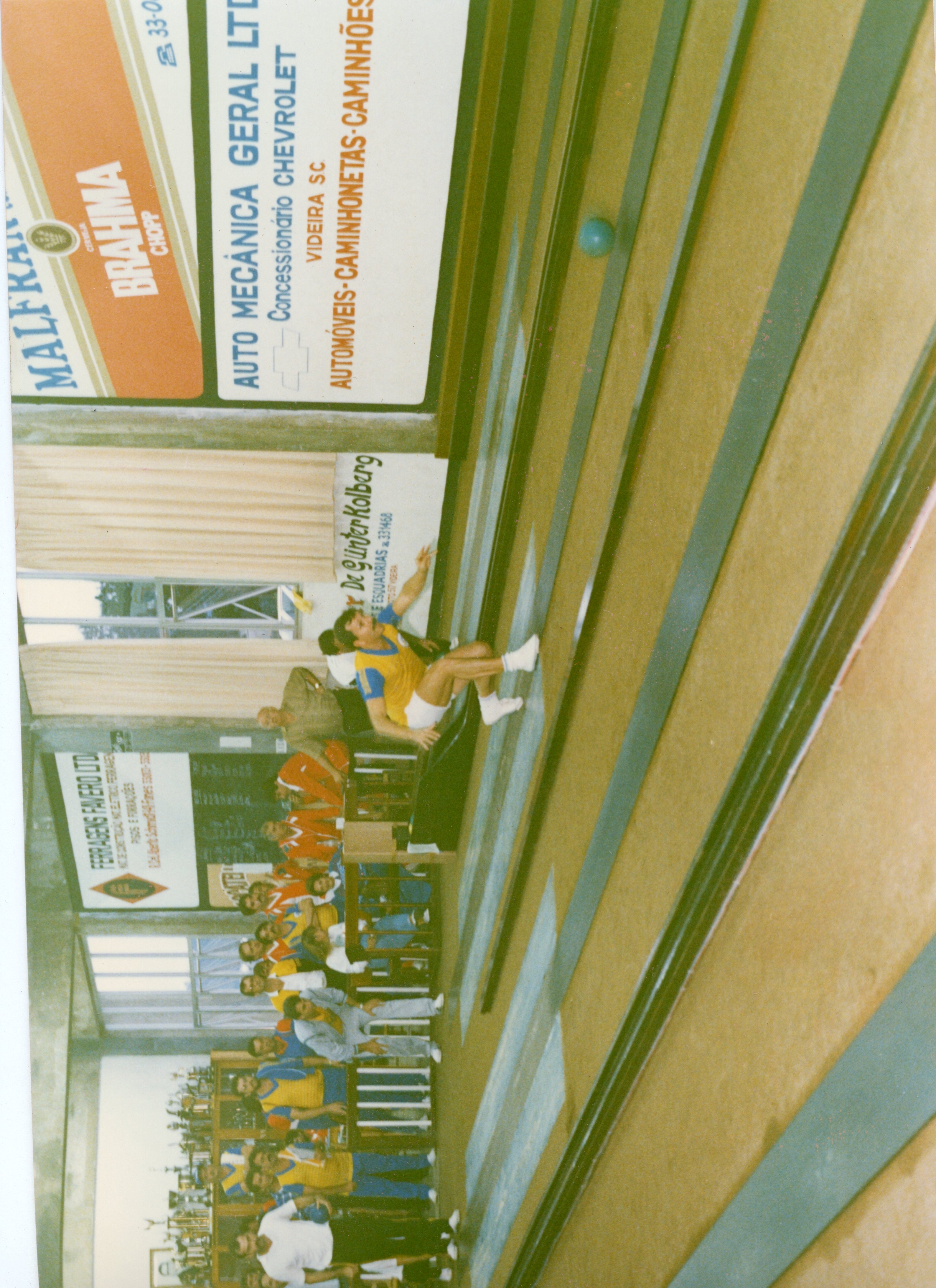 Uma fotografia de um torneio de bolão em um ambiente fechado, provavelmente em Videira, Santa Catarina, dado os anúncios locais nas paredes. O registro mostra um jogador em movimento na pista, enquanto outros participantes aguardam sentados em bancos, sugerindo uma atividade social e esportiva típica de clubes recreativos da região nas décadas de 1980 ou 1990.