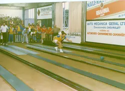 Uma fotografia de um torneio de bolão em um ambiente fechado, provavelmente em Videira, Santa Catarina, dado os anúncios locais nas paredes. O registro mostra um jogador em movimento na pista, enquanto outros participantes aguardam sentados em bancos, sugerindo uma atividade social e esportiva típica de clubes recreativos da região nas décadas de 1980 ou 1990.