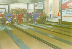 Uma fotografia de um grupo de homens praticando boliche em uma pista coberta, possivelmente em um clube social em Videira, Santa Catarina. O ambiente apresenta diversos anúncios comerciais nas paredes e uma estante repleta de troféus, sugerindo um contexto de competição esportiva amadora ou torneio local durante as décadas de 1980 ou 1990.