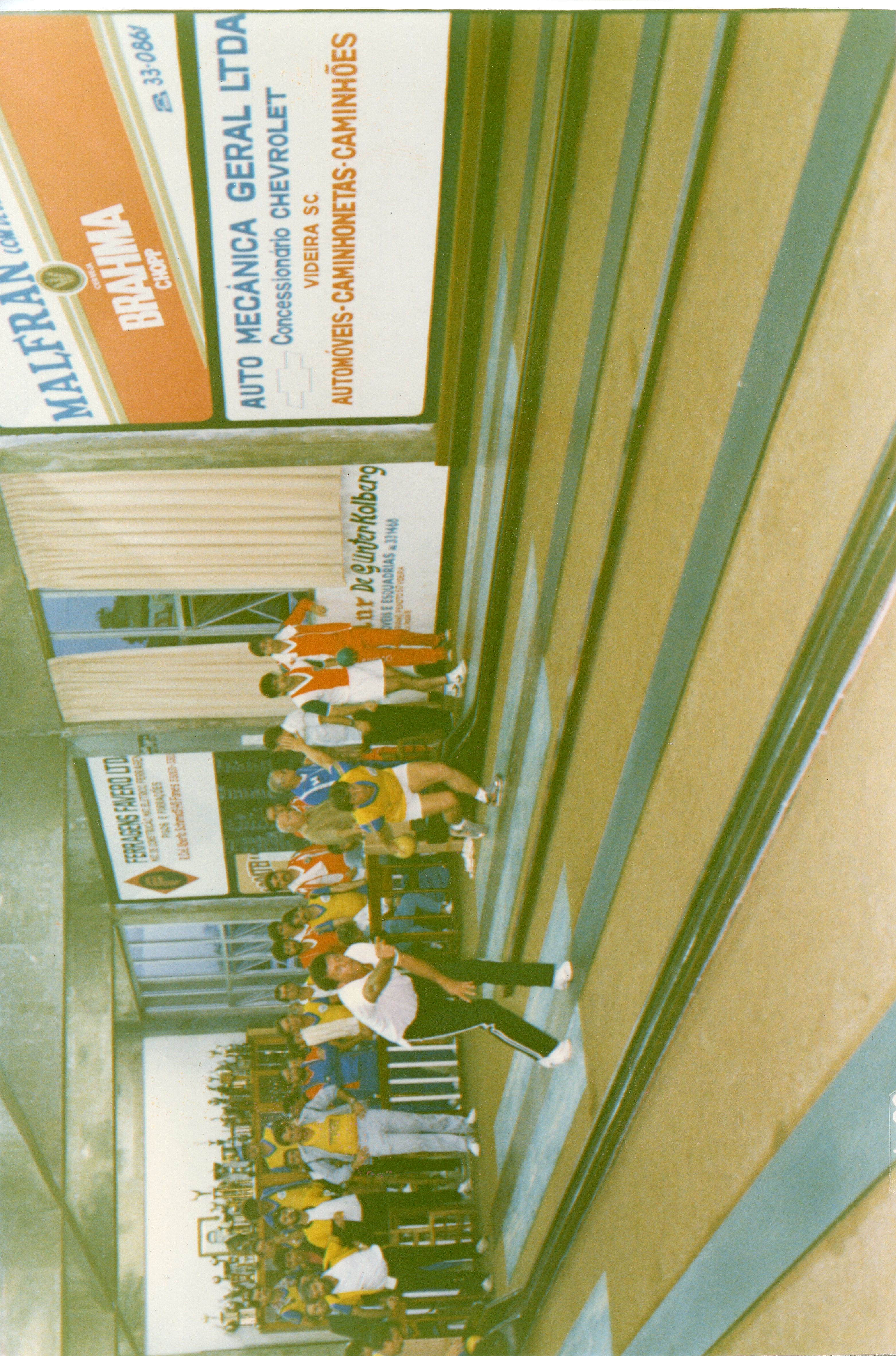 Uma fotografia de um grupo de pessoas praticando bolão em um ambiente fechado, provavelmente em Videira, Santa Catarina, dado o contexto dos anúncios locais. A imagem apresenta diversos jogadores em ação e troféus ao fundo, sugerindo um torneio ou atividade recreativa comum nas décadas de 1980 ou 1990.