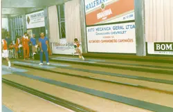 Uma fotografia de um evento esportivo, possivelmente um jogo de bolão, realizado em um ambiente fechado com diversas placas publicitárias locais de Videira, SC. A imagem mostra jogadores em ação e em espera, com vestimentas esportivas típicas de meados da década de 1980 ou 1990.