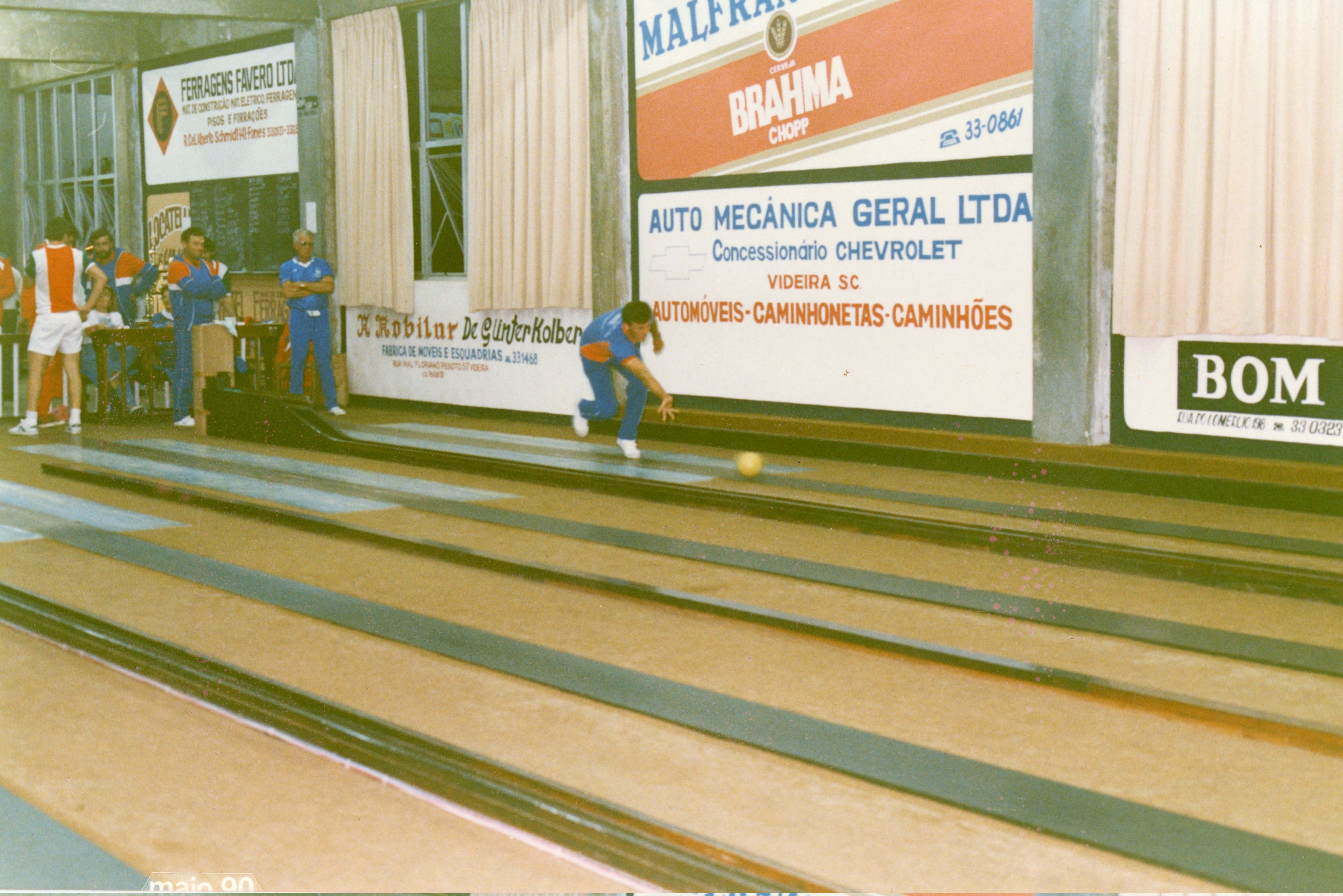 Uma fotografia colorida que registra uma partida de bolão em um clube social na cidade de Videira, Santa Catarina, datada de maio de 1990. O ambiente apresenta pistas de madeira e paredes decoradas com anúncios publicitários de empresas locais da época, enquanto um jogador realiza um lançamento.