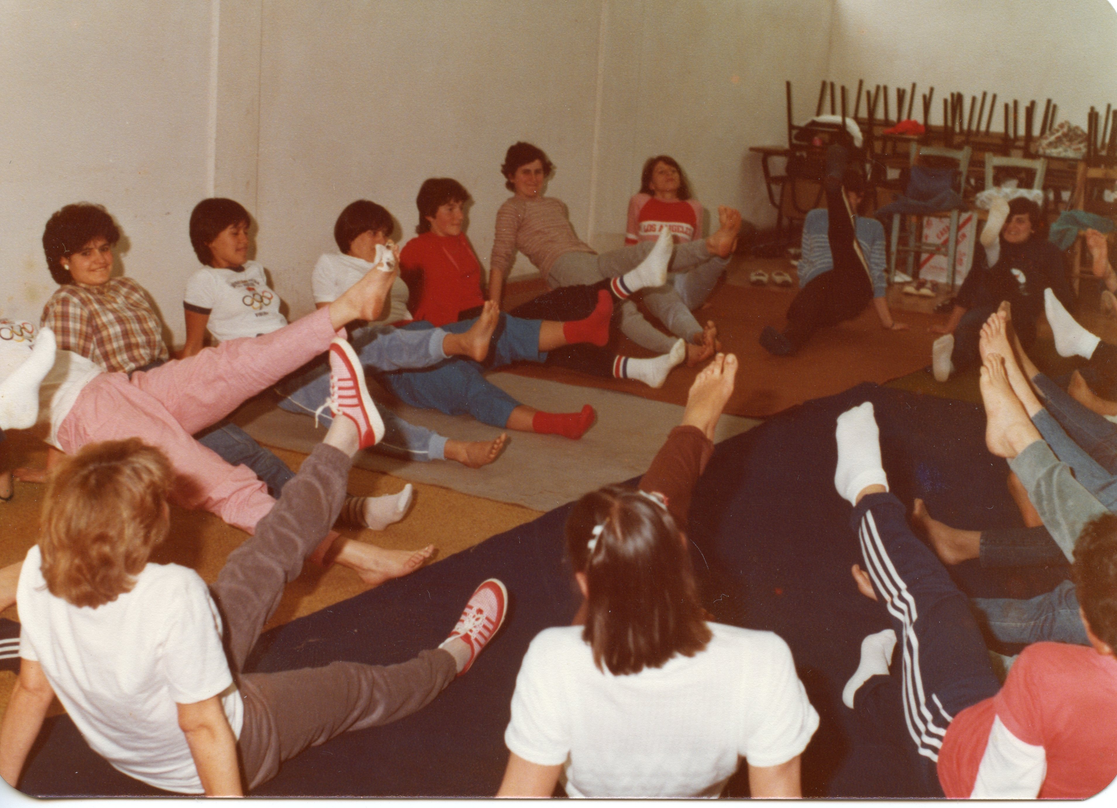 Uma fotografia colorida que retrata um grupo de jovens mulheres praticando exercícios físicos ou ginástica em um ambiente interno, possivelmente um salão comunitário ou escola. As participantes vestem roupas típicas da década de 1980, como calças de moletom e camisetas, e estão sentadas em colchonetes realizando movimentos de alongamento com as pernas elevadas.