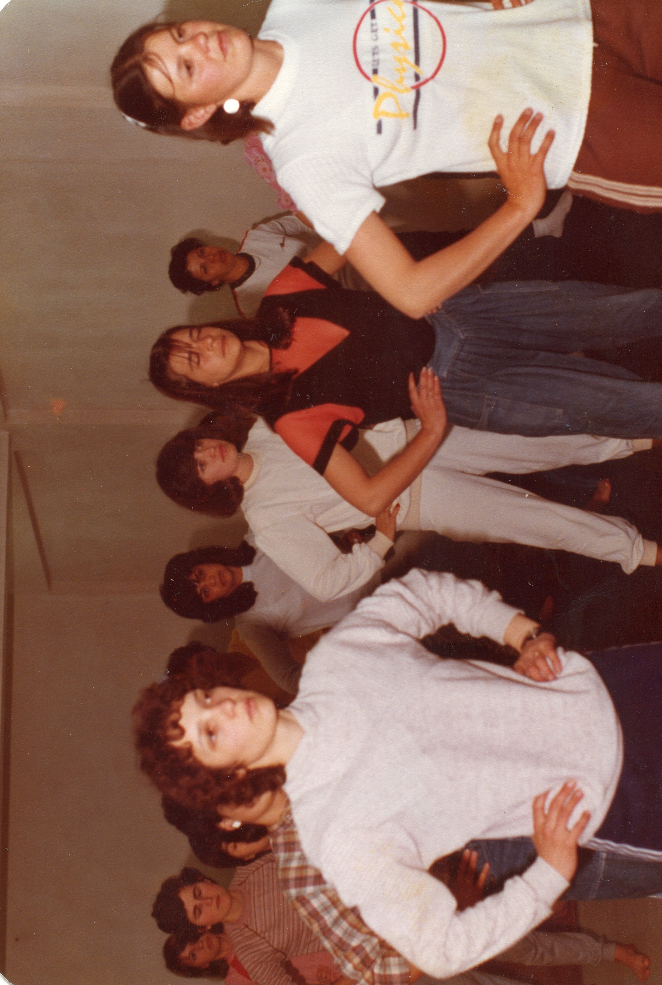 Uma fotografia colorida de época, provavelmente das décadas de 1980 ou 1990, mostrando um grupo de jovens mulheres em um ambiente interno, possivelmente uma escola ou centro comunitário. Elas vestem roupas casuais típicas da época, como moletons e camisetas, e parecem estar participando de uma atividade recreativa ou ensaio.