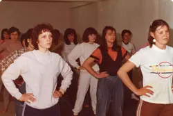Uma fotografia colorida de época, provavelmente das décadas de 1980 ou 1990, mostrando um grupo de jovens mulheres em um ambiente interno, possivelmente uma escola ou centro comunitário. Elas vestem roupas casuais típicas da época, como moletons e camisetas, e parecem estar participando de uma atividade recreativa ou ensaio.