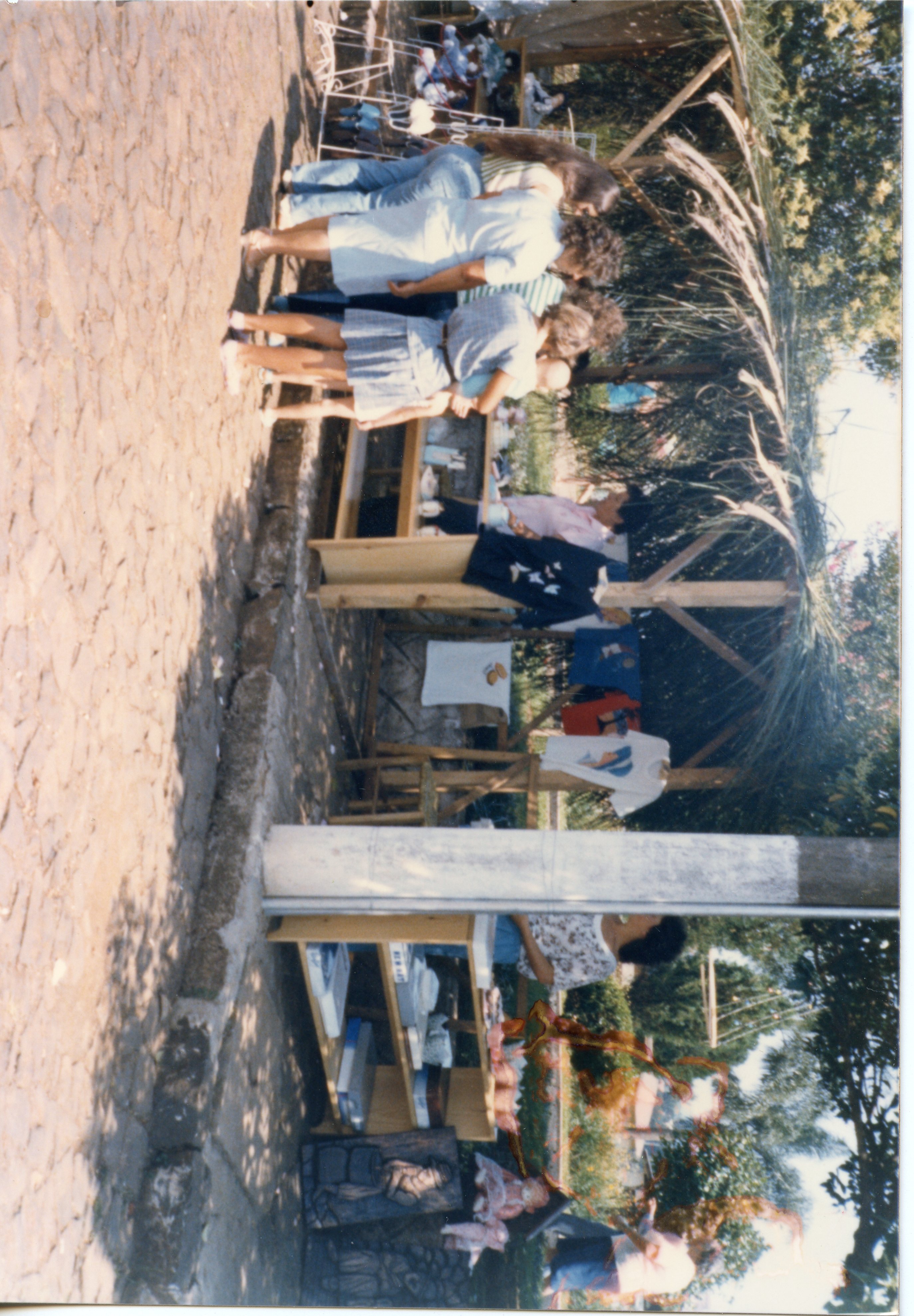 Uma fotografia colorida que mostra uma feira ou evento ao ar livre com barracas de madeira cobertas por palha. Pessoas estão reunidas examinando roupas e objetos expostos em bancadas, sugerindo um contexto de comércio local ou evento comunitário típico de pequenas cidades brasileiras nas décadas de 1980 ou 1990.