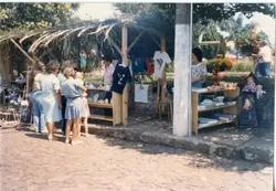 Uma fotografia colorida que mostra uma feira ou evento ao ar livre com barracas de madeira cobertas por palha. Pessoas estão reunidas examinando roupas e objetos expostos em bancadas, sugerindo um contexto de comércio local ou evento comunitário típico de pequenas cidades brasileiras nas décadas de 1980 ou 1990.