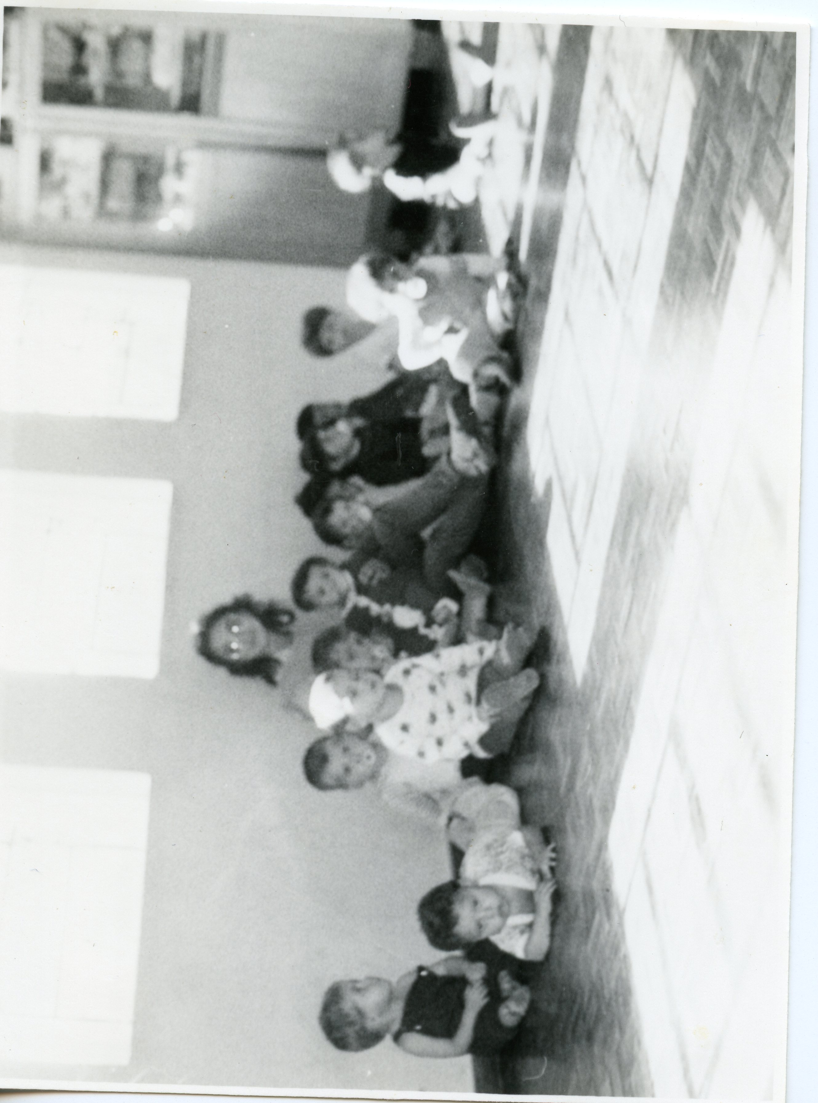Fotografia em preto e branco capturando um grupo de crianças sentadas no chão de uma sala, possivelmente em uma escola ou creche. A imagem, com estética típica de meados do século XX, mostra um ambiente interno simples com janelas laterais e piso de madeira, sugerindo uma atividade recreativa ou educacional.