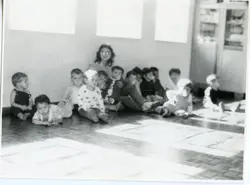 Fotografia em preto e branco capturando um grupo de crianças sentadas no chão de uma sala, possivelmente em uma escola ou creche. A imagem, com estética típica de meados do século XX, mostra um ambiente interno simples com janelas laterais e piso de madeira, sugerindo uma atividade recreativa ou educacional.