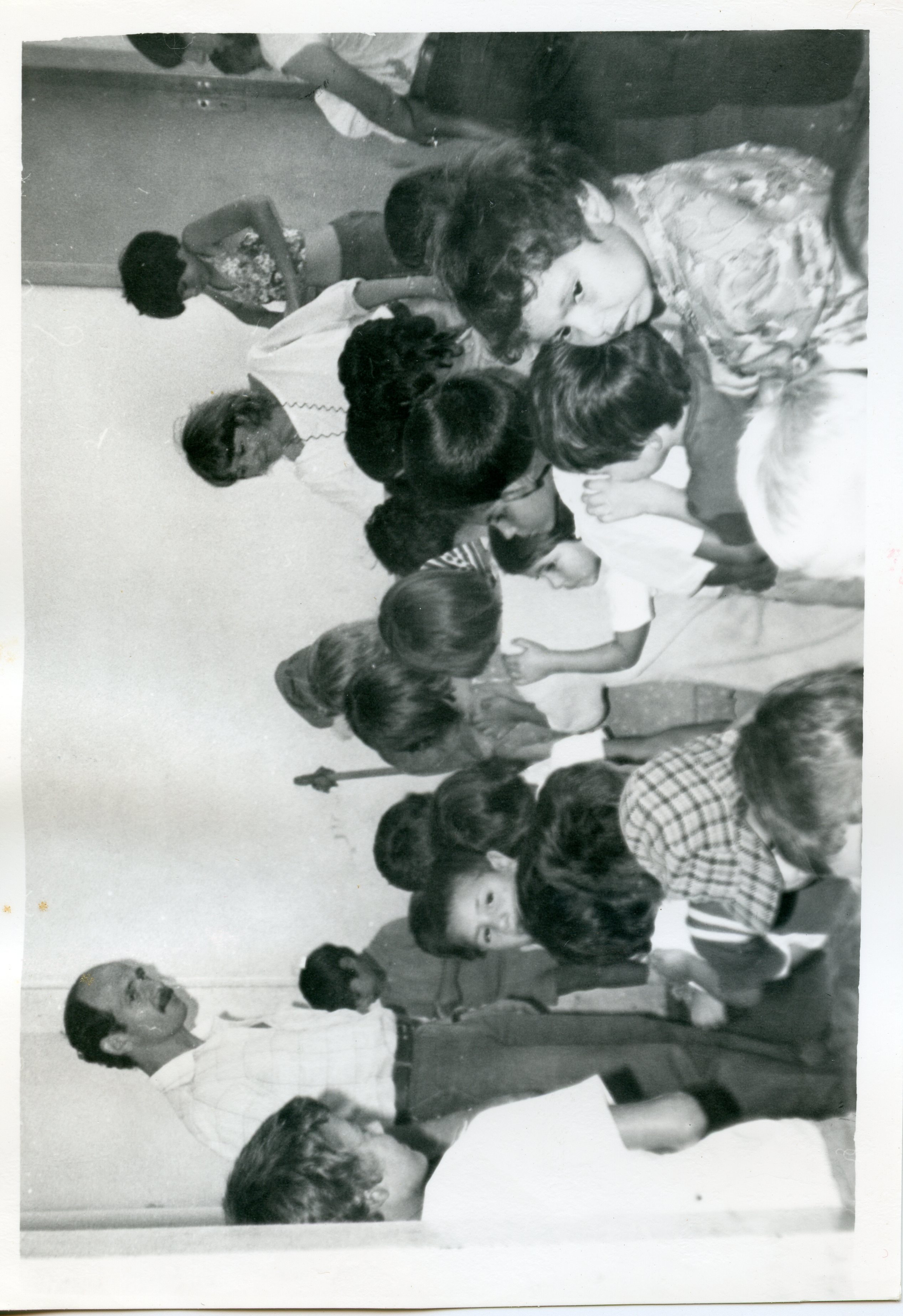 Fotografia em preto e branco que retrata um grupo de crianças reunidas em um ambiente interno, possivelmente uma sala de aula ou espaço comunitário. Um homem adulto, vestindo camisa social, observa o grupo, sugerindo uma atividade educativa ou recreativa típica de meados da década de 1970 ou 1980.