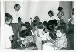 Fotografia em preto e branco que retrata um grupo de crianças reunidas em um ambiente interno, possivelmente uma sala de aula ou espaço comunitário. Um homem adulto, vestindo camisa social, observa o grupo, sugerindo uma atividade educativa ou recreativa típica de meados da década de 1970 ou 1980.