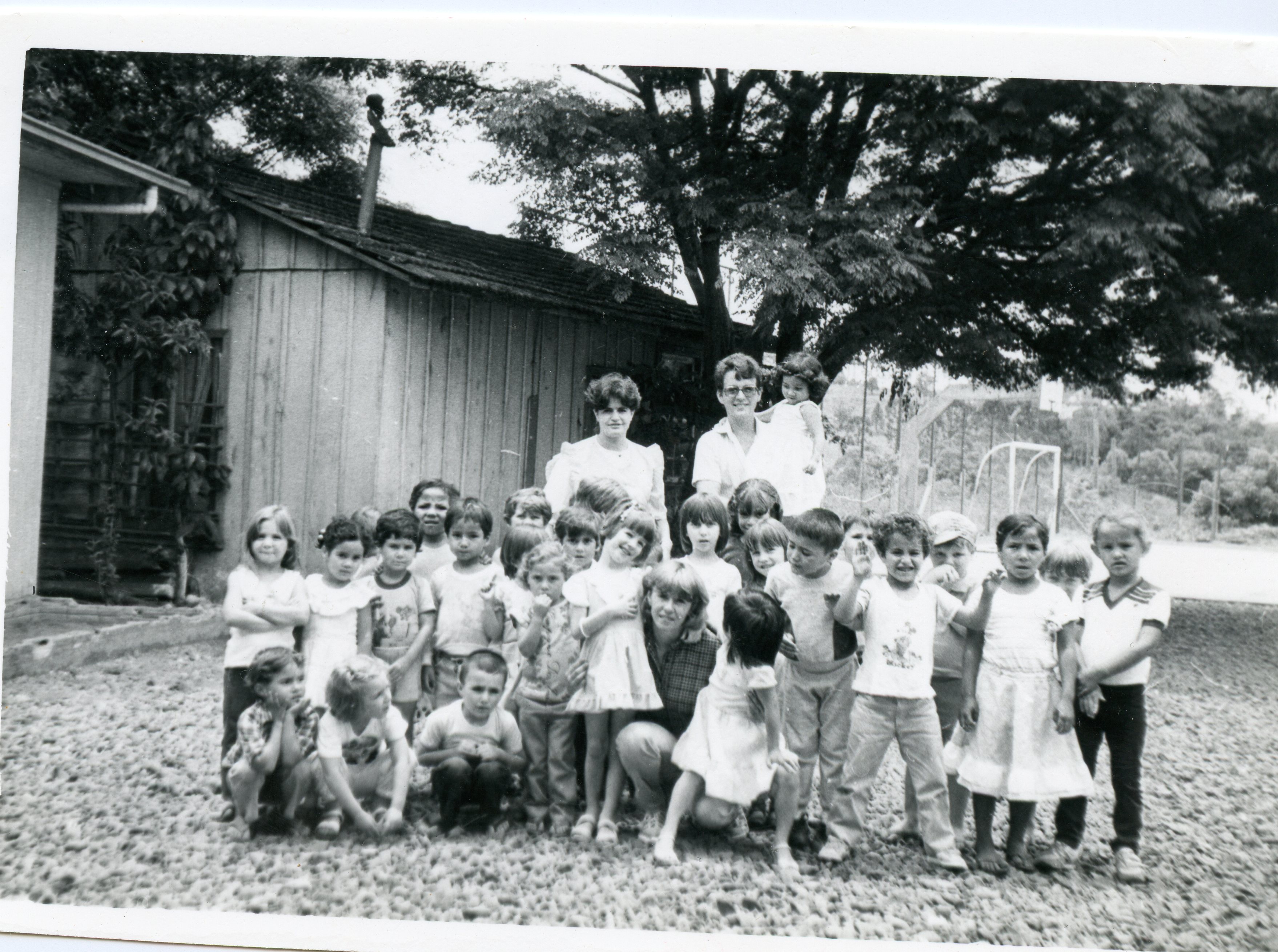 Fotografia em preto e branco de um grupo de crianças posando com duas adultas em frente a uma casa de madeira simples. A cena sugere uma atividade escolar ou recreativa em uma área rural ou suburbana, possivelmente na década de 1970 ou 1980.