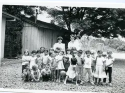 Fotografia em preto e branco de um grupo de crianças posando com duas adultas em frente a uma casa de madeira simples. A cena sugere uma atividade escolar ou recreativa em uma área rural ou suburbana, possivelmente na década de 1970 ou 1980.