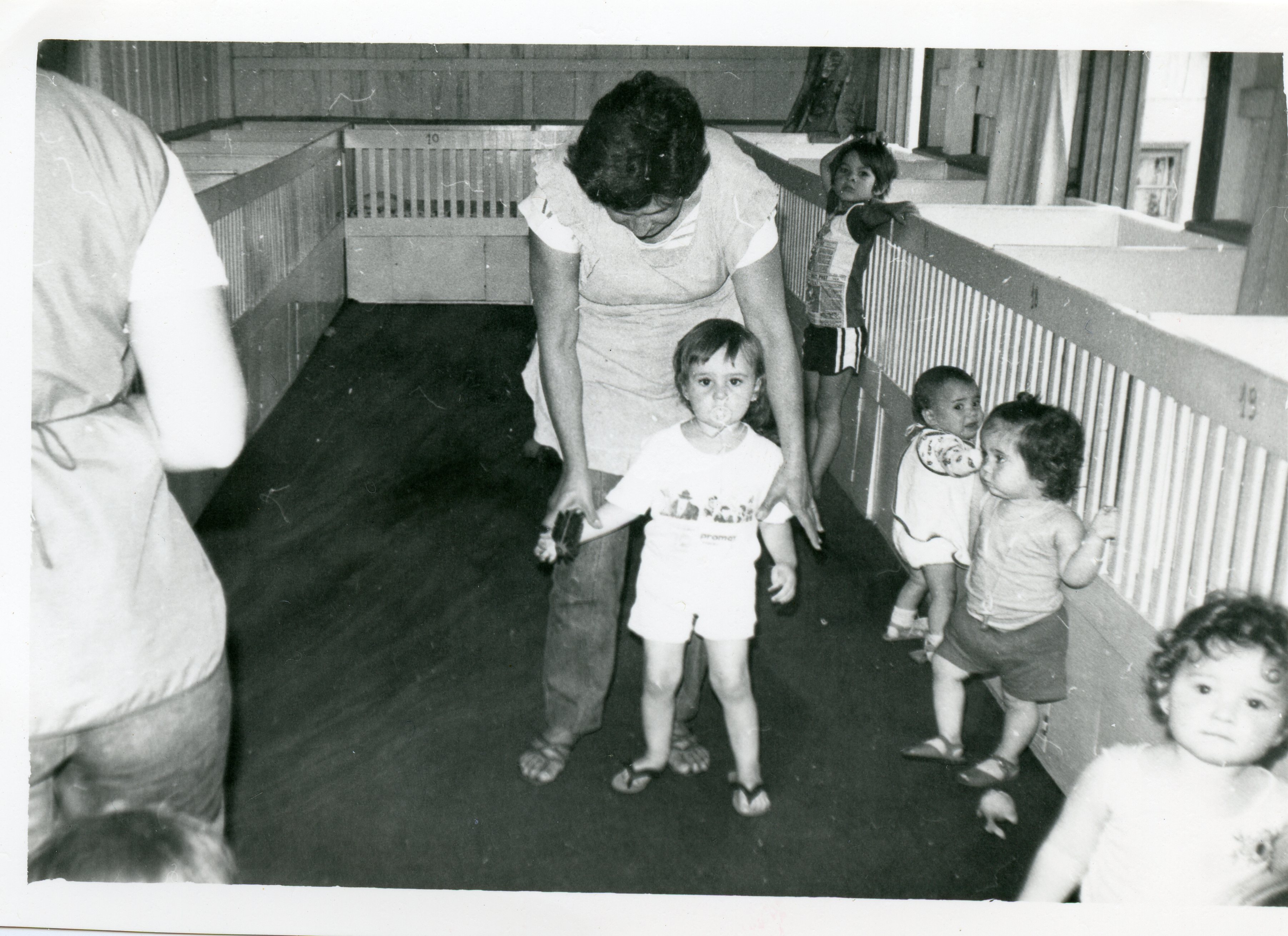 Fotografia em preto e branco de uma creche ou berçário, mostrando uma cuidadora auxiliando uma criança a caminhar entre cercadinhos de madeira. O ambiente sugere uma instituição de cuidado infantil, possivelmente datada da década de 1970 ou 1980, com crianças pequenas em um espaço coletivo.