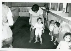 Fotografia em preto e branco de uma creche ou berçário, mostrando uma cuidadora auxiliando uma criança a caminhar entre cercadinhos de madeira. O ambiente sugere uma instituição de cuidado infantil, possivelmente datada da década de 1970 ou 1980, com crianças pequenas em um espaço coletivo.