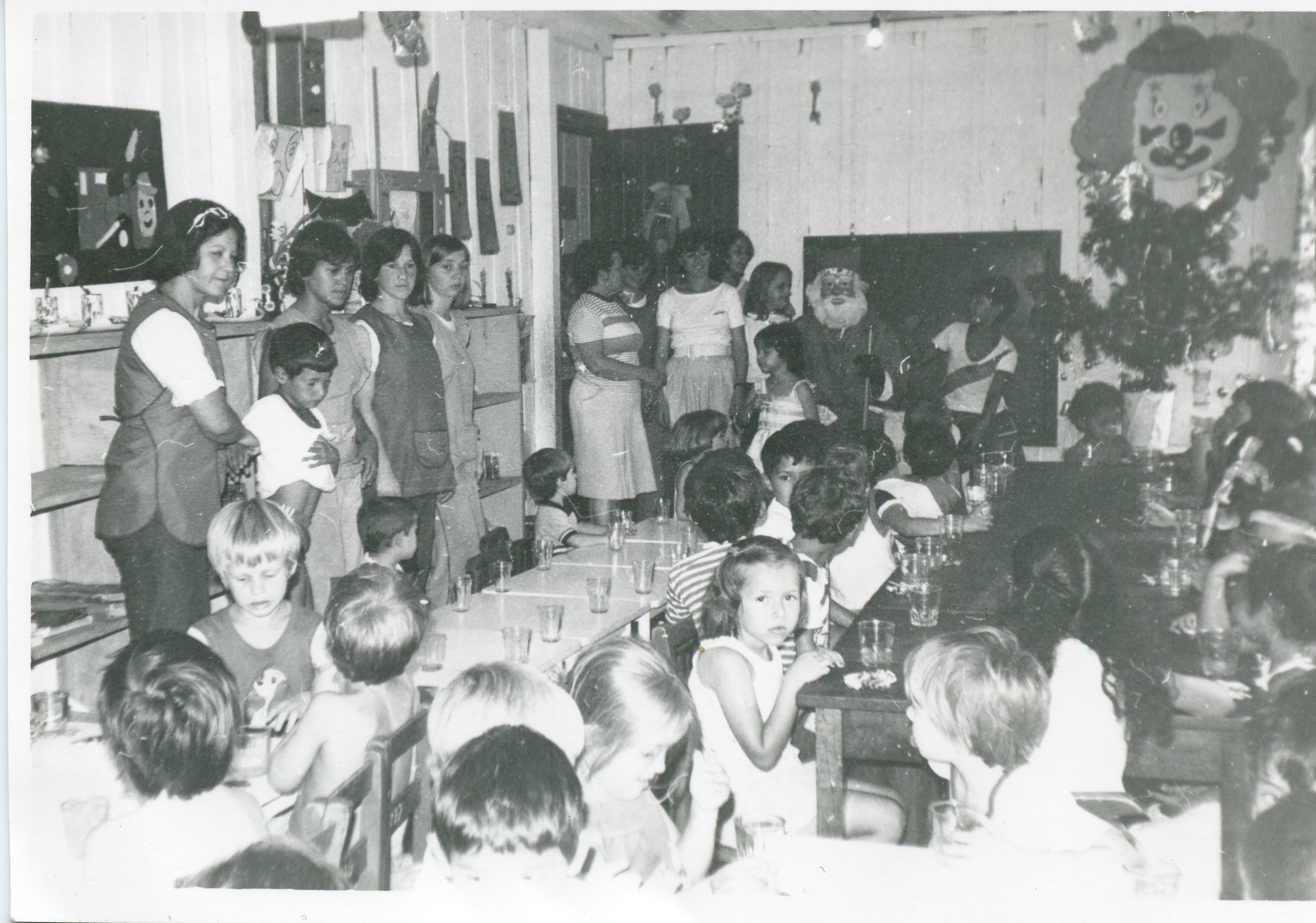 Uma fotografia em preto e branco que retrata uma celebração de Natal em uma escola ou centro comunitário, possivelmente nas décadas de 1970 ou 1980. Vê-se um grupo de crianças sentadas em mesas, adultos supervisionando e uma figura vestida de Papai Noel, com decoração temática de palhaço na parede.