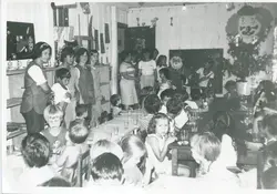 Uma fotografia em preto e branco que retrata uma celebração de Natal em uma escola ou centro comunitário, possivelmente nas décadas de 1970 ou 1980. Vê-se um grupo de crianças sentadas em mesas, adultos supervisionando e uma figura vestida de Papai Noel, com decoração temática de palhaço na parede.