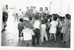 Fotografia em preto e branco capturando uma fila de crianças em um ambiente interno, possivelmente uma escola ou centro comunitário. As vestimentas e o estilo da imagem sugerem um contexto brasileiro de meados do século XX, possivelmente entre as décadas de 1960 e 1970.