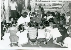 Uma fotografia em preto e branco que retrata uma celebração infantil, possivelmente uma festa de Natal em uma escola ou creche, com a presença de uma figura vestida de Papai Noel cercada por crianças sentadas no chão. A cena sugere um ambiente escolar ou comunitário típico de meados do século XX, com decoração simples e vestimentas casuais da época.