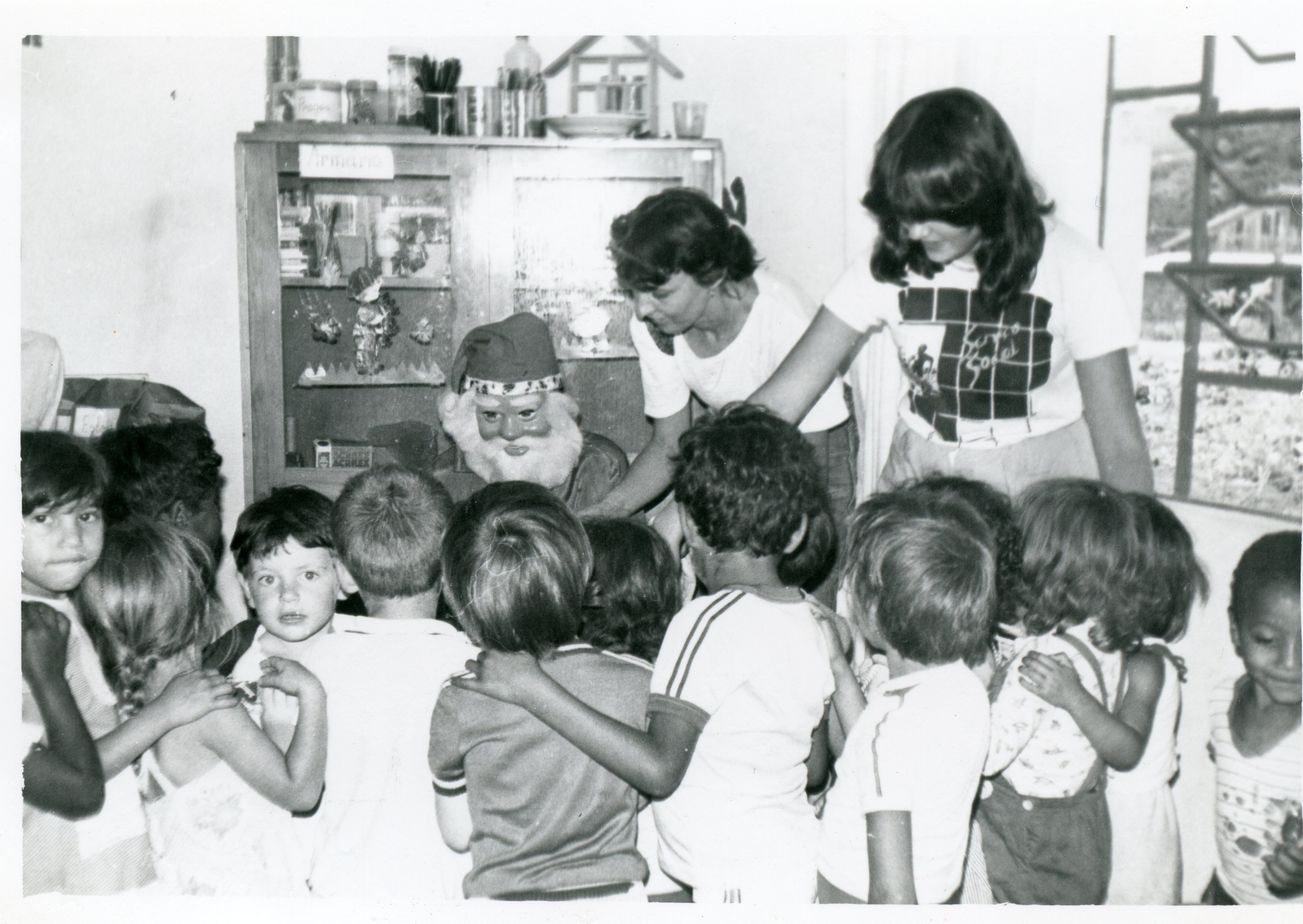 Fotografia em preto e branco retratando uma celebração natalina em um ambiente escolar ou comunitário, onde uma pessoa fantasiada de Papai Noel interage com um grupo de crianças. O cenário inclui um armário com objetos decorativos ao fundo e duas mulheres adultas auxiliando na organização, sugerindo uma atividade pedagógica ou social típica das décadas de 1970 ou 1980.