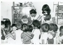 Fotografia em preto e branco retratando uma celebração natalina em um ambiente escolar ou comunitário, onde uma pessoa fantasiada de Papai Noel interage com um grupo de crianças. O cenário inclui um armário com objetos decorativos ao fundo e duas mulheres adultas auxiliando na organização, sugerindo uma atividade pedagógica ou social típica das décadas de 1970 ou 1980.