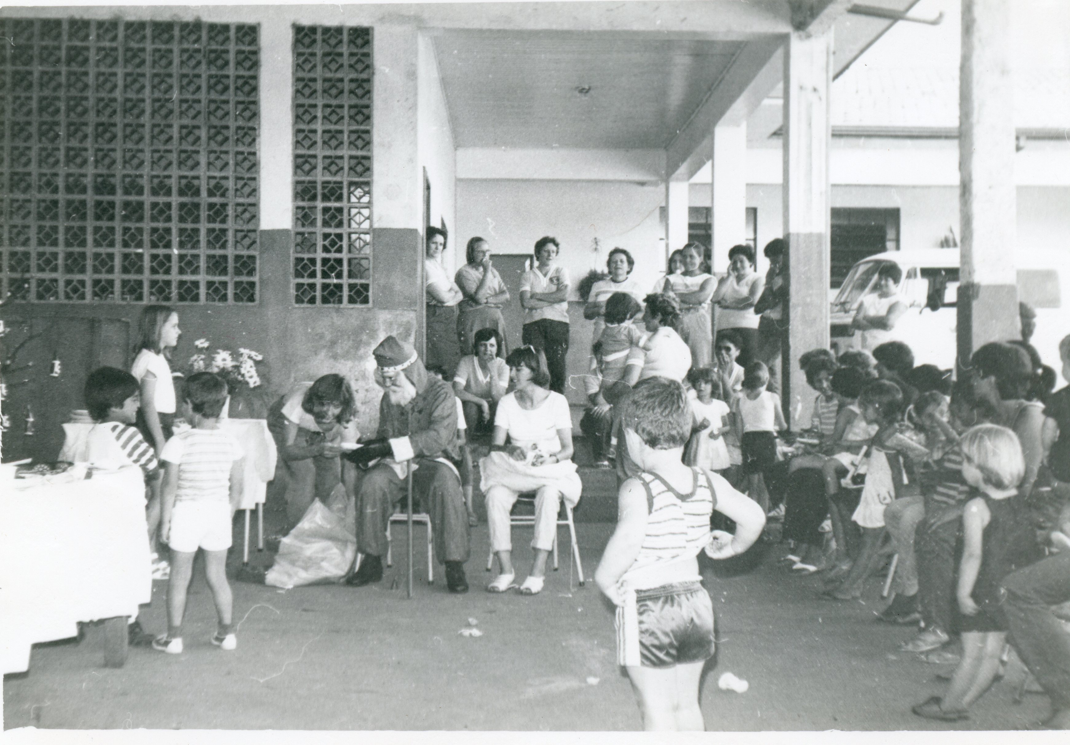 Uma fotografia em preto e branco que registra uma celebração de Natal com a presença de um Papai Noel, cercado por crianças e adultos em um ambiente que parece ser uma escola ou centro comunitário. A arquitetura com cobogós e a vestimenta das pessoas sugerem uma cena típica do cotidiano brasileiro entre as décadas de 1970 e 1980.