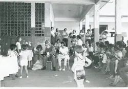 Uma fotografia em preto e branco que registra uma celebração de Natal com a presença de um Papai Noel, cercado por crianças e adultos em um ambiente que parece ser uma escola ou centro comunitário. A arquitetura com cobogós e a vestimenta das pessoas sugerem uma cena típica do cotidiano brasileiro entre as décadas de 1970 e 1980.